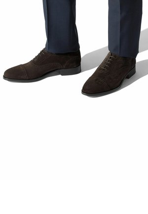 Brune suede sko med spids toe, detaljeret syning og en lav sort sål, parret med mørke bukser.