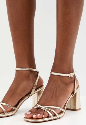 Voeten met metallic gouden sandalen met blokhak, dunne enkelbandjes en gekruiste tebandjes op een effen witte achtergrond.
