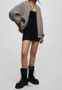 Chaqueta bomber de gran tamaño en color topo sobre un vestido mini negro fruncido, combinado con gruesas botas de combate negras con cordones, mostrando una variedad de texturas superpuestas.