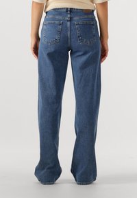 Személy magas derekú, egyenes szárú kék farmernadrágot és bézs színű, bordázott felsőt visel, háttal áll, kezei oldalt pihennek.