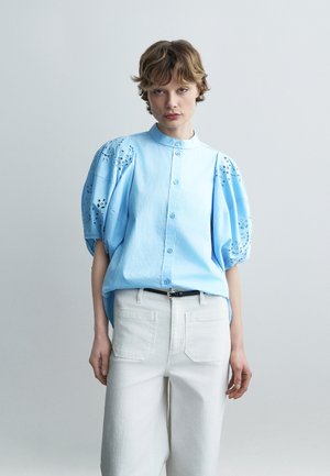 Young person wearing a light blue button-up blouse with puffed, lace-cut sleeves and white high-waisted pants with a black belt.