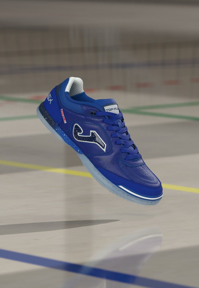 Niebieski sportowy but halowy z białą podeszwą i logo, unoszący się nad podłogą sali gimnastycznej z kolorowymi liniami boiska.