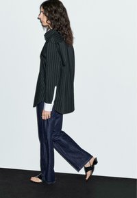 Donna con capelli mossi, indossa una camicia nera a righe sottili, jeans scuri a gamba larga e sandali neri con tacco aperto, mentre cammina davanti a un muro bianco. 