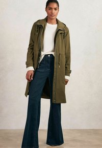 Imperméable vert olive avec une capuche, fermeture éclair à l'avant et poches latérales. Assorti avec un jean évasé noir et un pull blanc avec des poignets côtelés.
