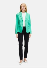 Světlý zelený blazer s dvěma předními kapsami, zapínáním na jeden knoflík a klopovými límci, nošený přes bílou košili a černé kalhoty.