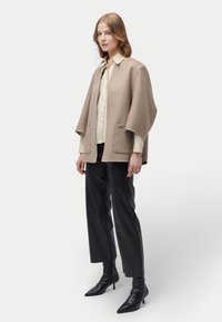 Frau steht und trägt eine beige übergroße Wolljacke über einem cremefarbenen Hemd, schwarze gekürzte Hosen und schwarze Stiefeletten mit spitzem Absatz.