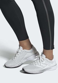 Zapatillas deportivas blancas con parte superior de malla texturizada, cierre de cordones y suela acolchada. Llevadas con leggings negros que tienen una franja lateral.