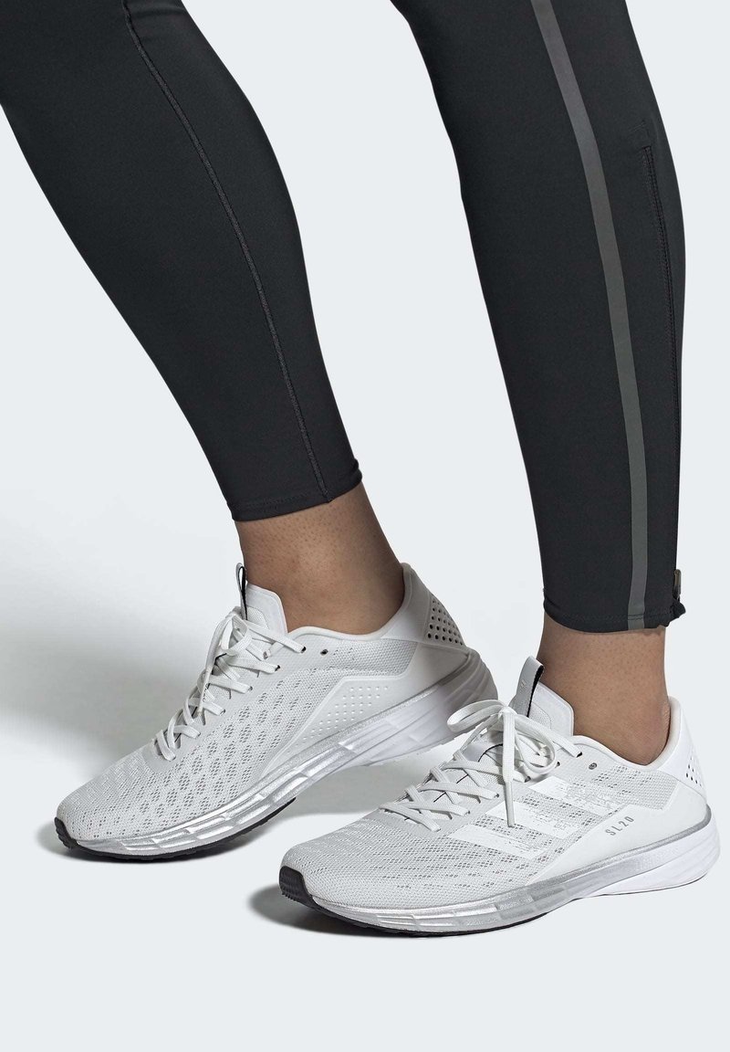 Zapatillas deportivas blancas con parte superior de malla texturizada, cierre de cordones y suela acolchada. Llevadas con leggings negros que tienen una franja lateral.