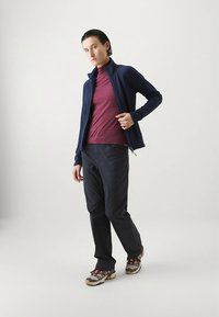 Jachetă bleumarin cu fermoar peste un pulover maro închis cu guler înalt, asortată cu pantaloni negri și adidași multicolori, pe un fundal alb simplu.