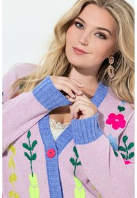 Jasnoróżowy sweter rozpinany z kolorowym kwiatowym haftem, niebieskim prążkowanym kołnierzem i jaskraworóżowym guzikiem. Miękka dzianina i wygodny krój.