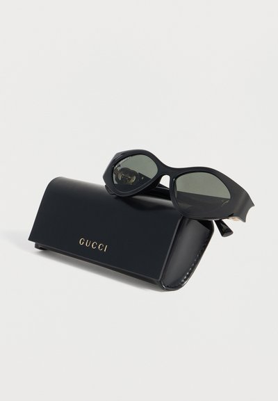 Μαύρα γυαλιά ηλίου σε σχήμα γατίσιου ματιού, που ξεκουράζονται σε μια μαύρη δερμάτινη θήκη Gucci με χρυσά γράμματα πάνω σε λευκό φόντο.