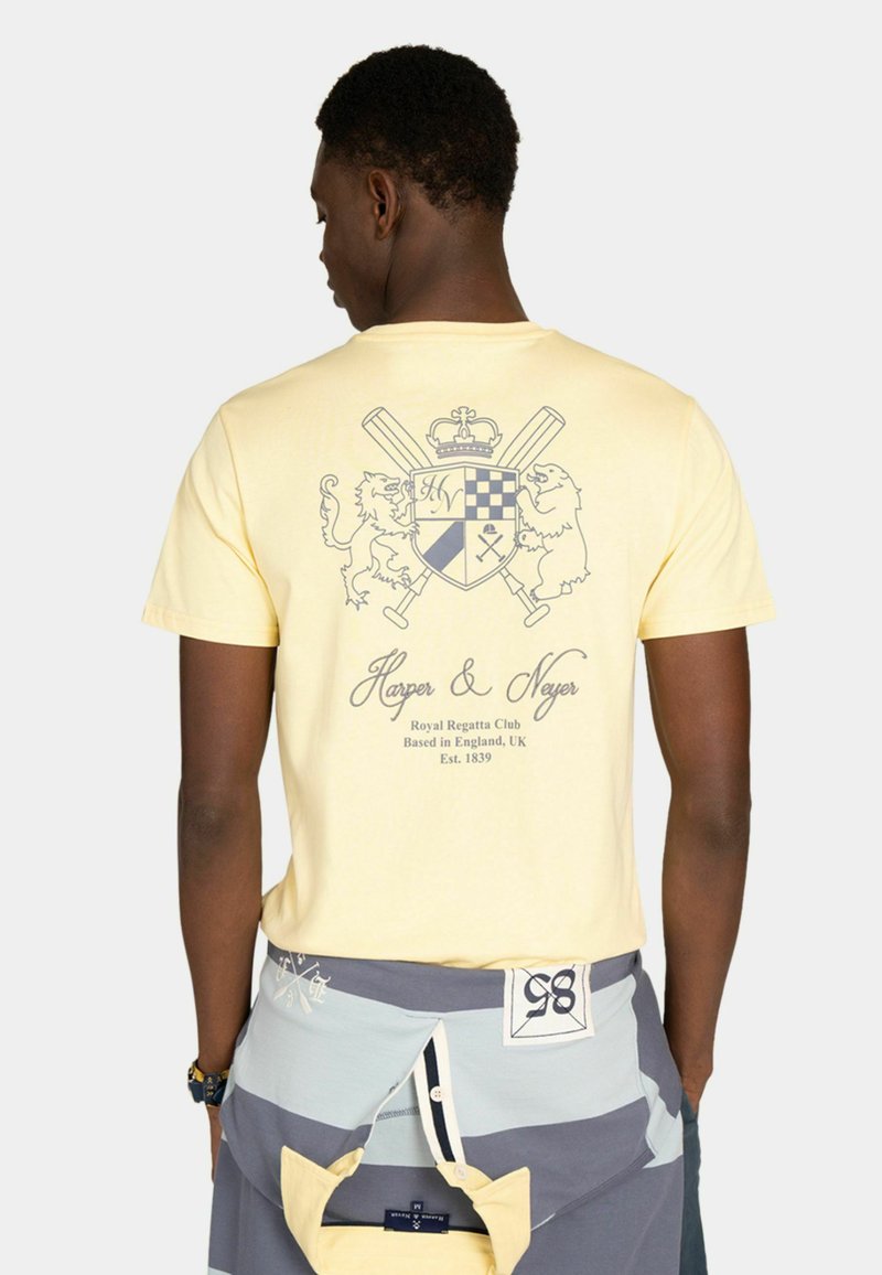 Homme portant un T-shirt jaune pâle avec un blason héraldique représentant un lion, un ours, des battes croisées et le texte « Harper & Neyer, Royal Regatta Club, England ».