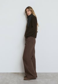 Brauner Strickpullover kombiniert mit weitem braunen Hosen. Ein dunkelgrau Schal ist um den Hals gelegt. Das Outfit zeichnet sich durch weiche Texturen und neutrale Farben aus.