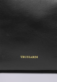 Czarna teksturowana skóra z wytłoczonym złotym logo „TRUSSARDI”. Gładkie wykończenie, prostokątny kształt, minimalistyczny design, podkreślający wysoką jakość materiału.