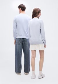 Δύο μοντέλα στέκονται πλάτη με πλάτη, φορώντας ανοιχτόχρωμα μπλε πουλόβερ· το ένα φορά φαρδιά τζιν και αθλητικά παπούτσια, ενώ το άλλο φούστα με πλισέ και λευκά αθλητικά παπούτσια.