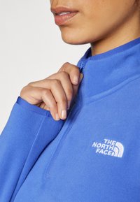 Plavi flis pulover s polu-zip ovratnikom, s izvezenim bijelim logotipom na prsima i glatkom, mekom teksturom.