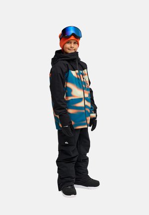 Kind trägt eine schwarz-blaue gemusterte Winterjacke, schwarze Schneehose, schwarze Handschuhe, orange Mütze und blaue Skibrille und steht vor weißem Hintergrund.