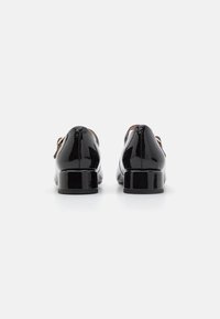 Chaussures en cuir verni noir avec un bout rond, un petit talon carré, et des accents décoratifs sur les côtés, présentant une surface lisse et réfléchissante.