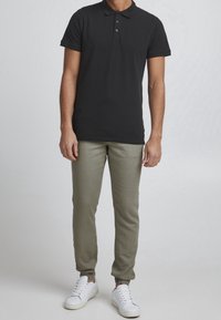 Czarna koszulka polo z trzema guzikami, zestawiona z oliwkowymi spodniami o wąskim kroju i białymi trampkami. Materiał wydaje się gładki, bez wzorów.