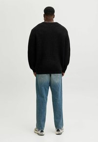Ein Mann steht mit dem Rücken zu uns, trägt einen schwarzen Pullover, ausgewaschene blaue Jeans und weiße Turnschuhe vor einem hellen, einfarbigen Hintergrund.