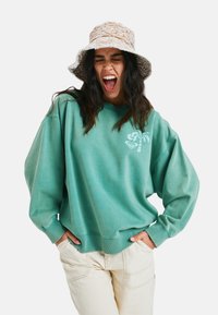 Jasnozielony, oversize'owy sweter z długimi rękawami i ściągaczami, z grafiką palmy na klatce piersiowej. W połączeniu z kremowymi spodniami.