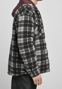 Veste en polaire à carreaux noirs et gris avec une fermeture éclair, une capuche bordeaux et deux poches latérales, présentant une texture douce et des manches longues.