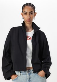 Giacca corta nera con colletto alto, zip frontale e vestibilità ampia, abbinata a una maglietta bianca e jeans azzurri. Tessuto morbido.