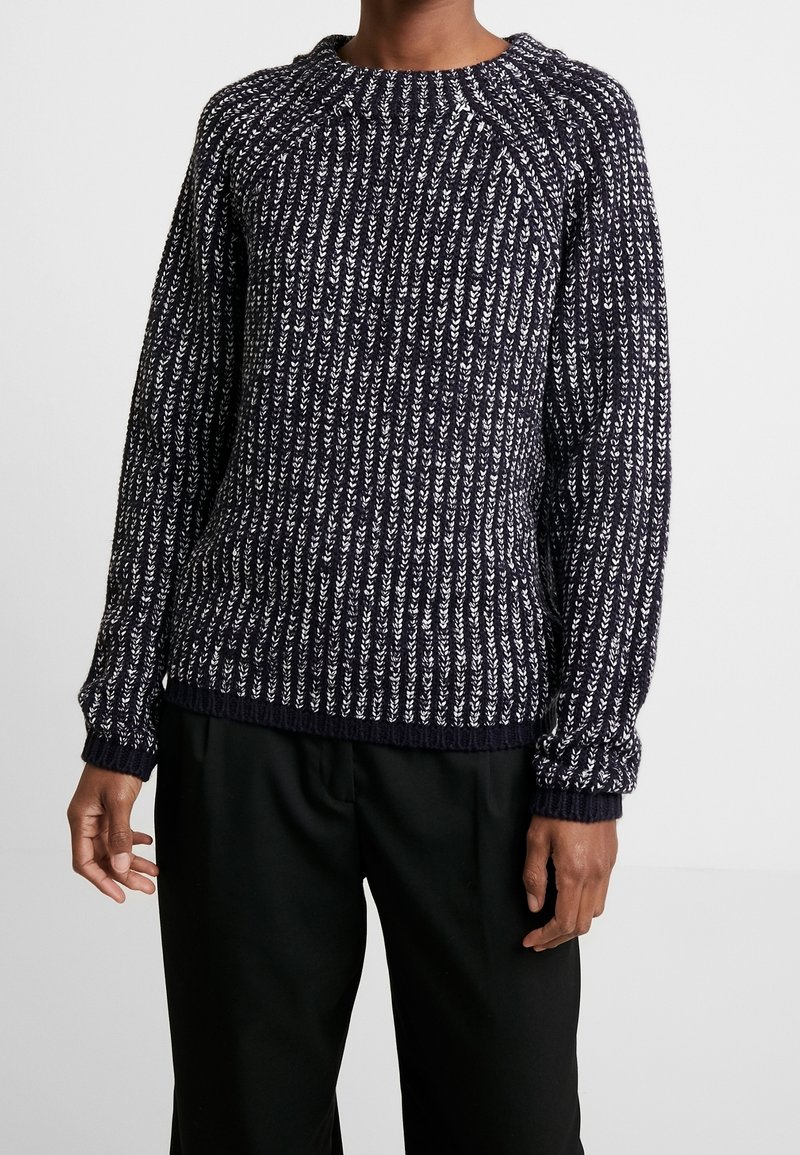 Marinblå stickad tröja med vita fläckar, strukturerad ribbad design, rundad halsringning och långa ärmar. I kombination med svarta byxor som syns nedanför.