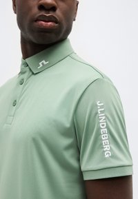 Άνδρας που φοράει ανοιχτοπράσινο πόλο με λογότυπο στον γιακά και το κείμενο "J.LINDEBERG" στο δεξί μανίκι, σε απλό φόντο.