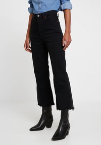 Jeans negro de tiro alto con pierna recta, dobladillo desflecado y sutilezas en la costura de los bolsillos, combinados con botas negras de tacón y una camisa de mezclilla.