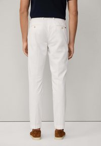 Pantaloni bianchi con vestibilità affusolata, tasche posteriori con dettagli in oro e polsini elasticizzati. Indossati con scarpe di camoscio marroni.