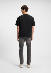 Fiatal férfi áll háttal, fekete pólót, szürke farmert és bézs sportcipőt viselve, egyszerű világos háttér előtt.