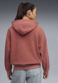 Oversized bluza z kapturem w kolorze przykurzonego różu, wykonana z teksturowanej tkaniny, z prążkowanymi mankietami, opadającymi ramionami i kieszenią kangurką. Nosi się ją z jasnoniebieskimi dżinsami.