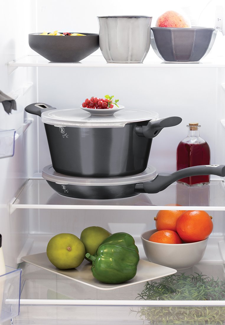 Deux casseroles noires avec des couvercles empilées dans un réfrigérateur, des raisins rouges sur une assiette, une bouteille de liquide rouge, des bols avec des fruits et des légumes verts.