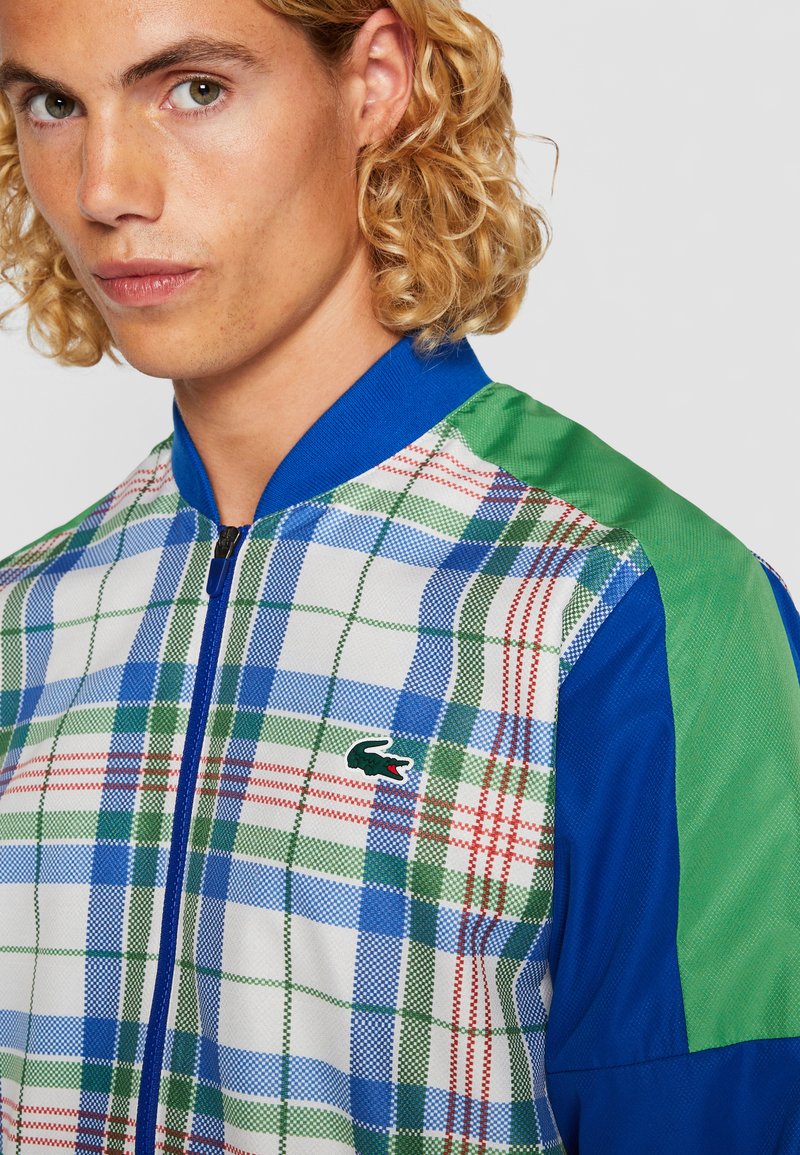 Veste bomber colorée avec un motif à carreaux bleu, vert et rouge, dotée d'un col bleu uni et d'accents d'épaule verts. Logo Lacoste inclus.