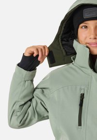 Jasnozielona wodoodporna kurtka z zapinana na zamek kieszenią na piersi, regulowanym kołnierzem i czarnymi akcentami. Wnętrze jest ciemne z teksturowanym wykończeniem.