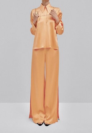 Satin two-piece outfit in soft orange, featuring a collared long-sleeve shirt and wide-leg pants with side slits. Black open-toe shoes.