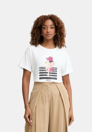 Donna con capelli ricci indossa una t-shirt bianca con grafica di cappello rosa e pantaloni beige a pieghe, a vita alta e gamba larga, in piedi davanti a uno sfondo uniforme.