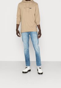 Beige huvtröja med lång ärm och text, ljusblå jeans, samt vita skor med svarta snören, framför en enfärgad bakgrund.