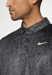 Černé polo tričko s texturovaným květinovým vzorem, se třemi knoflíky a bílým logem Nike na hrudi. Měkká tkanina a límeček.