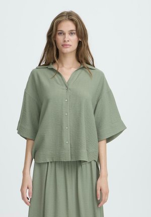 Zelená, textúrovaná blúzka s krátkym rukávom a špicatým golierom v kombinácii s prúdnou sukňou, čo vytvára súladný outfit.