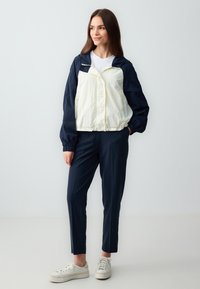 Lichte jas in crème en marineblauw, met een capuchon, elastische manchetten en een ontspannen pasvorm. Gepaard met marineblauwe broek en witte sneakers.
