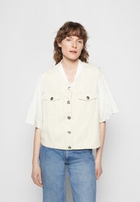 Colete de ganga bege com decote em V, dois bolsos frontais e botões prateados, combinado com uma blusa branca com franzidos e padrões de ilhós.