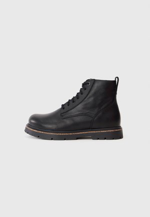 Bottine en cuir noir avec tige texturée, devant à lacets, languette à l'arrière, semelle épaisse en caoutchouc avec bordure beige, sur fond gris clair.