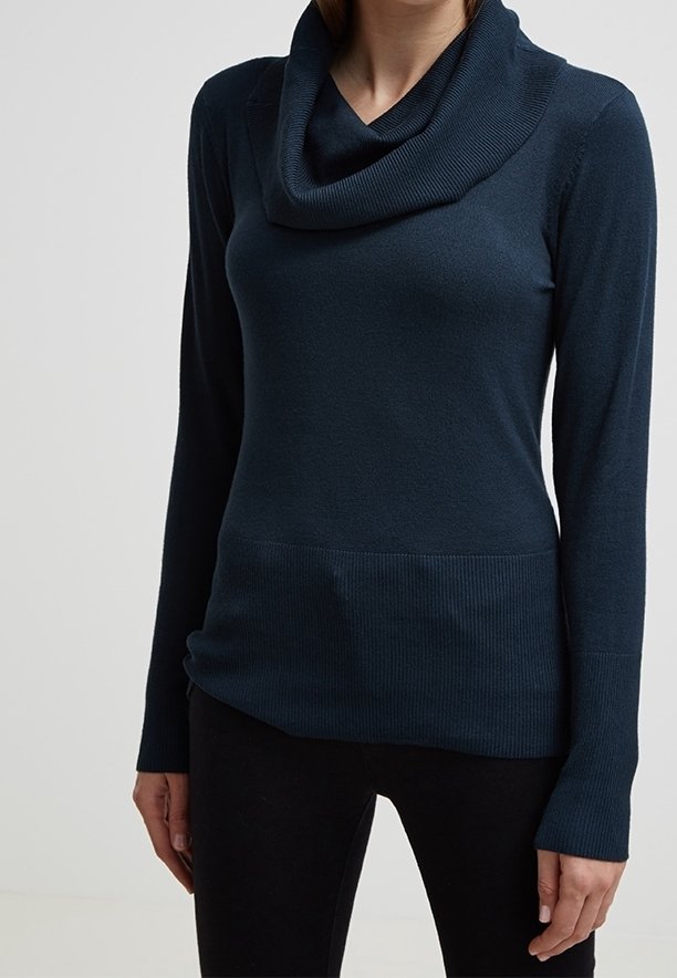 Femme portant un pull bleu foncé à manches longues avec col bénitier et ourlet côtelé, ainsi qu’un pantalon noir, debout devant un fond clair uni.