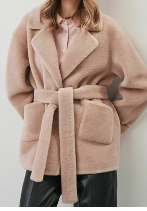 Kvinde iført beige, blød frakke med bælte over lyserød skjorte og sorte læderbukser, stående op ad en ensfarvet væg.