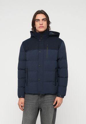 Jeune homme portant une veste matelassée bleu marine avec capuche, debout devant un fond clair uni.