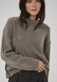 Hellbrauner Strickpullover mit hohem Hals, gerippter Textur, überschnittenen Schultern und breiten Bündchen, kombiniert mit dunklen Hosen.