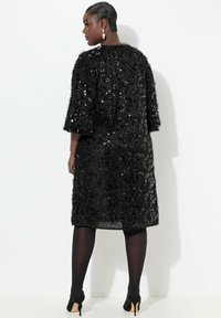Schwarzes Kleid aus strukturiertem Stoff, verziert mit Pailletten, mit drei Viertelärmeln und einem knielangen Saum, getragen mit schwarzen Strumpfhosen und Absatzschuhen.