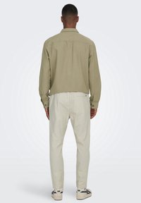 Olivfarbenes, langärmliges Hemd mit Knopfleiste und Rückenpasse; kombiniert mit hellbeigen Hosen und weißen Sneakers mit schwarzen Akzenten.